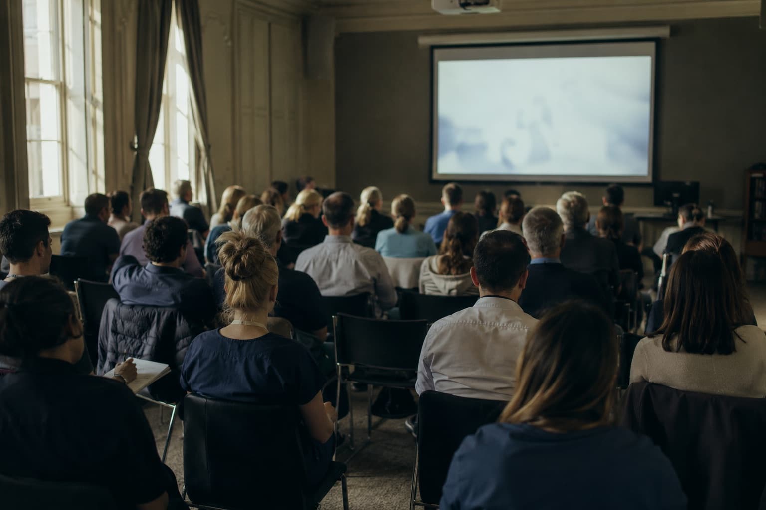A seminar room seen from the back, rows of seated people facing a glowing screen.